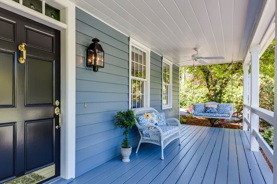 Front porch of a blue house with a dark front door, white wicker chairs, and a porch swing